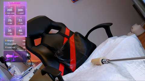 Video of a gamer's setup featuring a black, ergonomic gaming chair with red accents, a white keyboard, and a gaming mouse on a white desk. A digital overlay displays game statistics in purple text.
