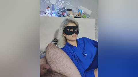 Media: A video shows a woman with platinum blonde hair wearing a black mask, blue shirt, and holding a man's leg. Background features a cluttered shelf with toys and decorations.