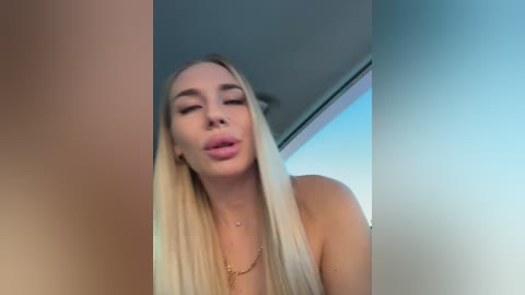 Video of a topless, blonde woman with long hair, full lips, and closed eyes, standing indoors with a blurred background.