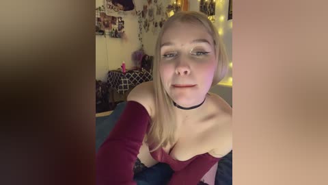 A video of a blonde, fair-skinned woman in a red off-shoulder top, with a black choker, lying on a bed with a blue blanket. Background shows a cozy room with posters, fairy lights, and a purple pillow.