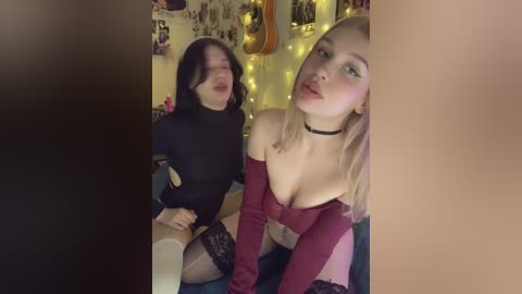 Video of two young women with fair skin and blonde hair, one in black lingerie and the other in a red top, sitting on a bed with string lights and posters in the background.