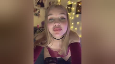 Video of a young blonde woman with fair skin, wearing a black choker and a red top, sitting in a cozy room with warm, yellow fairy lights in the background.