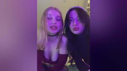 Video of two women with long hair, one blonde and one brunette, in a dark room with purple lighting. They are wearing off-shoulder dresses and have playful expressions, making pouty faces.