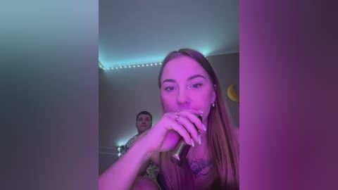 Media: Video of a young woman with long brown hair, wearing a floral top, holding a microphone, standing in a dimly lit room with purple lighting, visible ceiling lights, and a man in the background.
