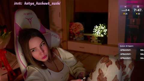 Media: Video of a young woman with light skin and straight brown hair, wearing a white top, seated in a pink gaming chair in a dimly lit room.