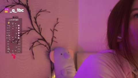 Video of a woman with long brown hair, wearing a white top, in a dimly lit room with purple lighting. She's positioned beside a wall with a black tree branch drawing, and a small white plush toy.