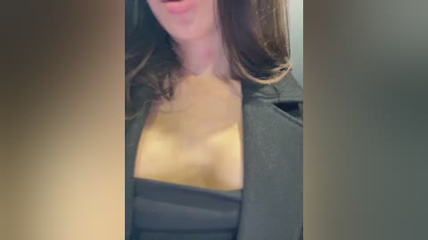 Media: Video of a light-skinned woman with shoulder-length brown hair, wearing a black blazer over a low-cut, revealing black top, exposing cleavage. She has a confident, slightly open-mouthed expression. The background is blurred, suggesting an indoor setting.
