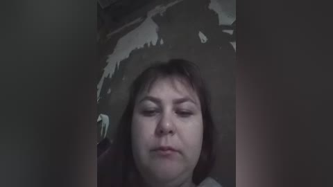 Video of a woman with short brown hair, wearing a gray shirt, lying on her back, eyes closed, in a dimly lit room with a shadowy figure in the background.