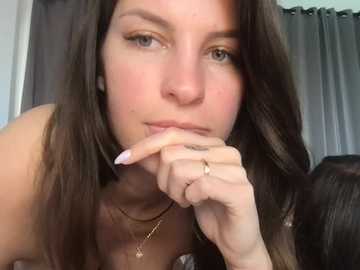 Video of a young woman with long brown hair, light skin, and a serene expression. She is leaning forward with her chin resting on her hand, wearing a delicate necklace. The background features dark curtains.