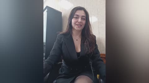 Video of a young woman with long, dark hair, wearing a black blazer and black skirt, sitting in a wooden chair against a beige wall, smiling softly.