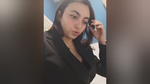 Video of a young woman with long dark hair, wearing a black blazer, applying lipstick in a bathroom with a blue wall and white door.