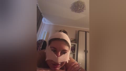 A video shows a woman wearing a pink, knitted face mask, smiling, in a dimly lit room with beige walls, a chandelier, and a mirror.