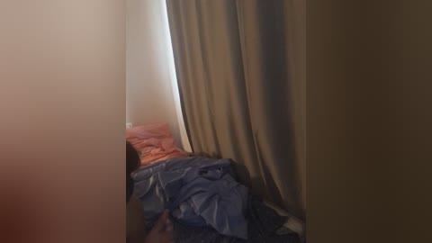 Video of a small, dimly lit room with beige walls and a bed covered in blue and orange sheets. The room appears cramped, with shadows and a sense of disorder.