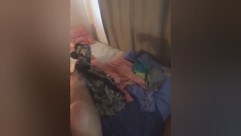 A video of a small, light-colored cat lying on a messy bed with a mix of pink, blue, and grey blankets. The cat's fur appears short and white with a few black patches.