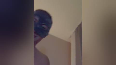 Video of a man with a dark mask covering his face, standing in a dimly lit room with beige walls. His figure is partially obscured by shadows.