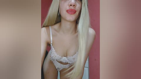 Video of a fair-skinned, blonde woman with red lipstick, wearing a lacy white bra, standing against a red wall, with a blurred background.