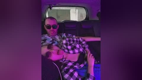 Video of a man and woman in a car, both wearing sunglasses, with purple lighting. The man is in a checkered shirt, and the woman is in a black top.