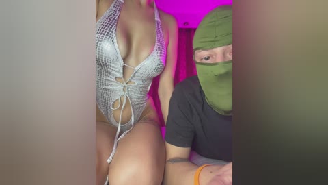 Video of a woman with light skin, wearing a revealing, silver mesh halter swimsuit, and a man in a green mask, seated in a dimly lit room with purple lighting.