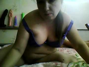 Video of a woman with light skin, wearing a purple bra, sitting on a bed with floral sheets, surrounded by various sex toys.