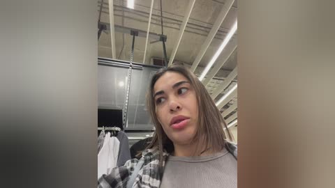 Media: Video of a young woman with straight, shoulder-length brown hair, wearing a gray sweater and checkered flannel, standing in a modern, industrial warehouse with exposed beams and fluorescent lights.