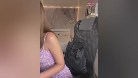 Media: Video of a woman in a light purple dress with a floral pattern, sitting on a bed with a grey backpack and a beige blanket. Background includes a white dresser and a mural with a glowing string light border.