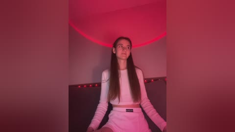 Media: Video of a young woman with long brown hair, wearing a white crop top and shorts, sitting on a dark couch under red lighting.