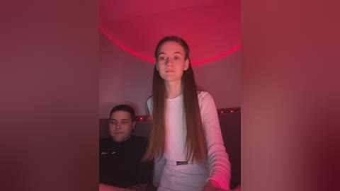 Media: Video of two young women, one with long straight brown hair, wearing a white long-sleeve top and light blue jeans, and the other with short dark hair, wearing a black top, against a red-lit background.