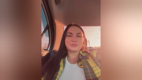 Video of a woman with long dark hair, wearing a yellow plaid shirt over a white T-shirt, inside a car with sunlight streaming through the window.