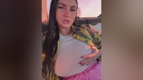 Video of a woman with long black hair, wearing a yellow and black patterned shirt, white top, and pink pants, seated on a dark couch, her right hand covering her chest.