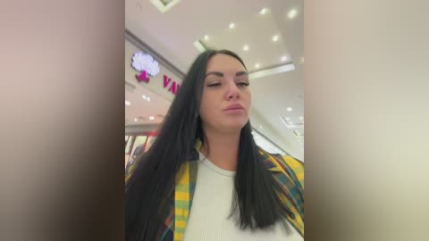 Video of a young woman with long, straight black hair and fair skin, wearing a white top and a plaid shirt, standing in a brightly lit, modern shopping mall.