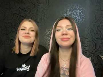 Video of two young women with light skin and straight hair, one with pink highlights and the other with dark brown hair. Both wear black T-shirts and pink hoodies. Background features intricate black wallpaper.