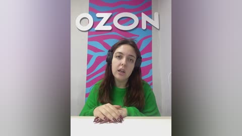 Video of a young woman with long brown hair and fair skin, wearing a green sweater, sitting at a white desk in a studio with a blue and pink zebra-striped backdrop.