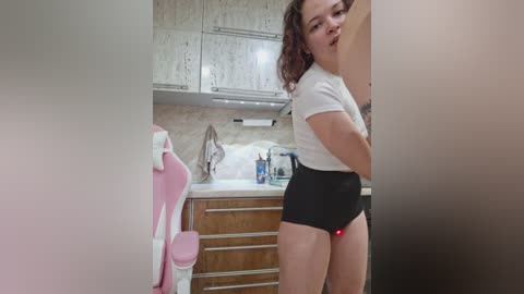 Media: A video of a curvy, fair-skinned woman with curly brown hair in a white top and black shorts, standing in a modern kitchen with beige cabinets, a sink, and pink chair.