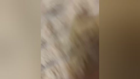 A blurred video features a person with light skin and indistinguishable facial features, likely wearing a light-colored shirt, standing against a beige wall with a slightly darker background. The image is out of focus, making details indiscernible.