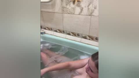 Media: Video of a woman with short brown hair, partially submerged in a bathtub filled with bubbles, her breasts visible, surrounded by beige and brown tiled walls.