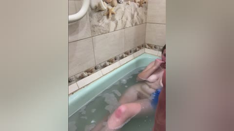 A video of a nude person with pale skin, small breasts, and a blue towel, submerged in a bathtub filled with bubbles, against a beige-tiled background.