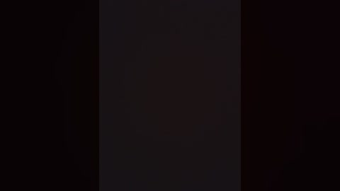 A dark, shadowy video of a tall, slender woman standing in the middle, wearing a black dress and black stockings. The background is mostly black, with faint, indistinct shapes and textures.