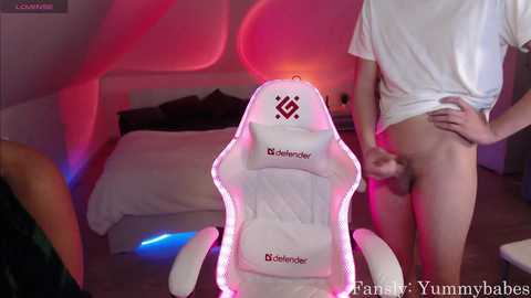 Media: A video of a white gaming chair with neon lights, a person pulling down their white shirt to reveal their bare backside in a dimly lit bedroom with a bed.