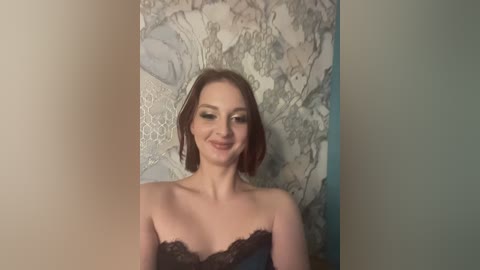 Media: Video of a smiling, fair-skinned woman with shoulder-length auburn hair, wearing a black strapless lace dress, standing in front of a textured, marble-like wall with intricate patterns.