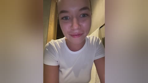 Media: A video of a young woman with light skin and blue eyes, wearing a white t-shirt, standing in a hallway with beige walls and a carpeted floor.