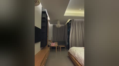 Media: Video of a modern, minimalist bedroom with a wooden bed, beige curtains, and a chandelier. A flat-screen TV is mounted on the wall, and a small table with chairs is situated near the bed. The room is lit with soft, ambient light.
