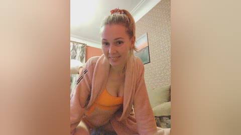 Media: A video of a smiling, fair-skinned woman with blonde hair tied in a ponytail, wearing a pink robe over an orange bra, sitting on a beige couch in a room with floral wallpaper.
