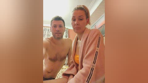 Media: A video of a shirtless, muscular white man with short brown hair and a woman with light skin and auburn hair in a ponytail, wearing a pink jacket and a yellow top, sitting closely together in a dimly lit room.