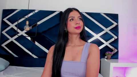 Video of a young woman with long black hair, wearing a light purple tank top, standing against a dark blue, diamond-patterned wall, with a modern bedside table and a purple lamp in the background.