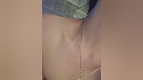 Video of a close-up of a person's bare shoulder, showing a delicate gold chain necklace. The skin is light-toned, and the background is blurred with a neutral beige hue.