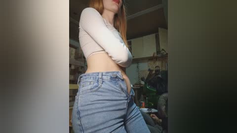 A video of a slim, fair-skinned woman with long, straight red hair, wearing a cropped white ribbed top and high-waisted blue jeans, posing in a cozy, cluttered living room with wooden cabinets and a green sofa.