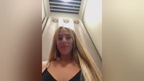 A video of a young Caucasian woman with long, straight blonde hair, wearing a black strapless top, standing in a narrow, dimly lit hallway with beige walls and a white ceiling with a single light fixture.