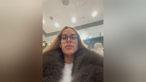 Video of a woman with long blonde hair, wearing black-framed glasses and a dark fur coat, seated indoors with a blurred background of chairs and a ceiling.