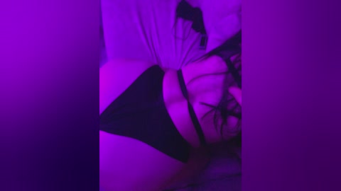 A video with purple lighting features a person in black lingerie, their face obscured, lying on a bed with white sheets. The image is blurred, emphasizing a sensual, intimate atmosphere.
