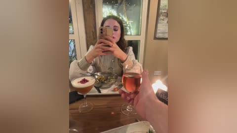 Video of a woman with long brown hair, wearing a light sweater, taking a selfie in a cozy, dimly-lit room with a wooden table and two wine glasses.